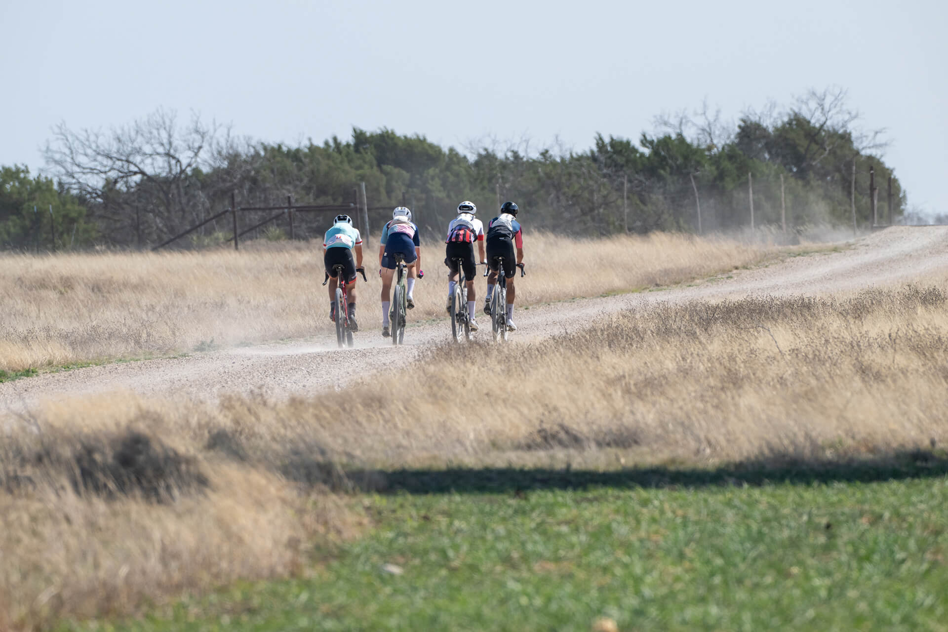 Five cyclists ride on a dusty gravel road with dry grass on either side, under a clear blue sky, conveying a sense of adventure and camaraderie.