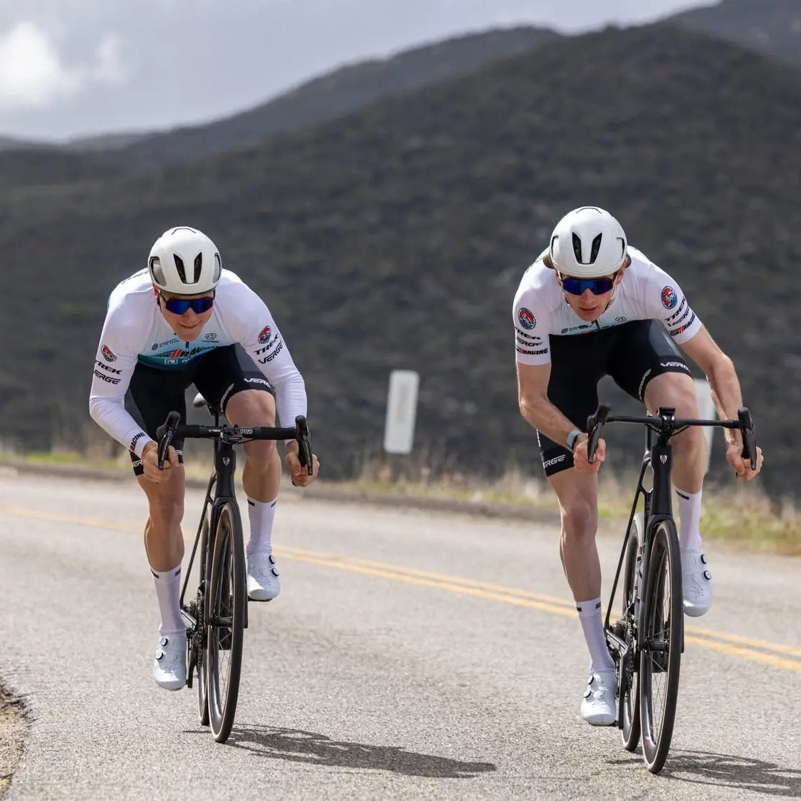 Two cyclists wearing white uniforms and helmets race down a winding road against a backdrop of rugged hills and cloudy skies, conveying speed and focus.