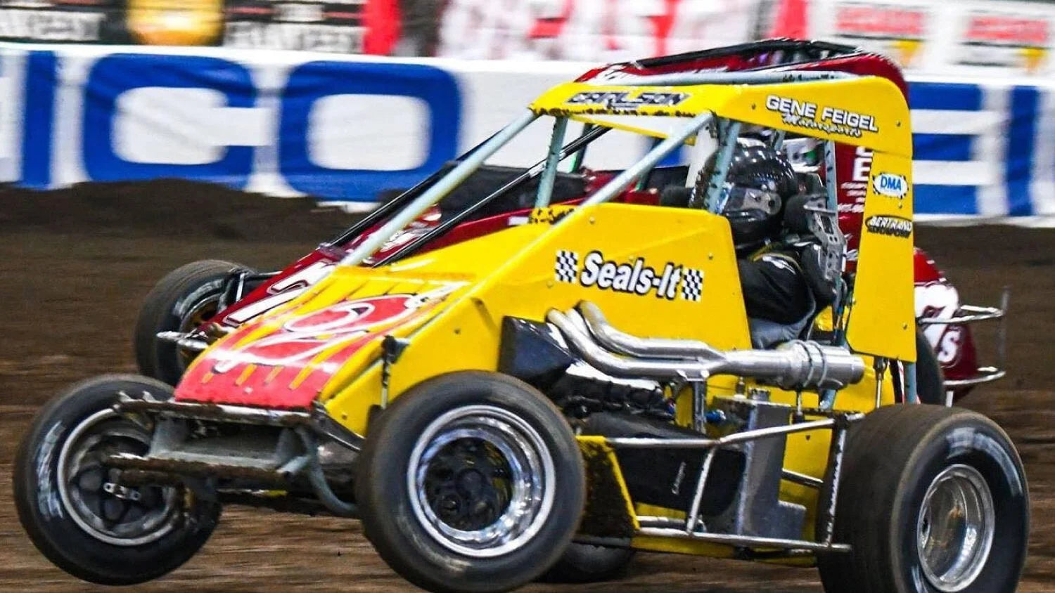 Two brightly-colored sprint cars race on a dirt track, kicking up dust. The cars are close, showcasing a tense, competitive atmosphere.