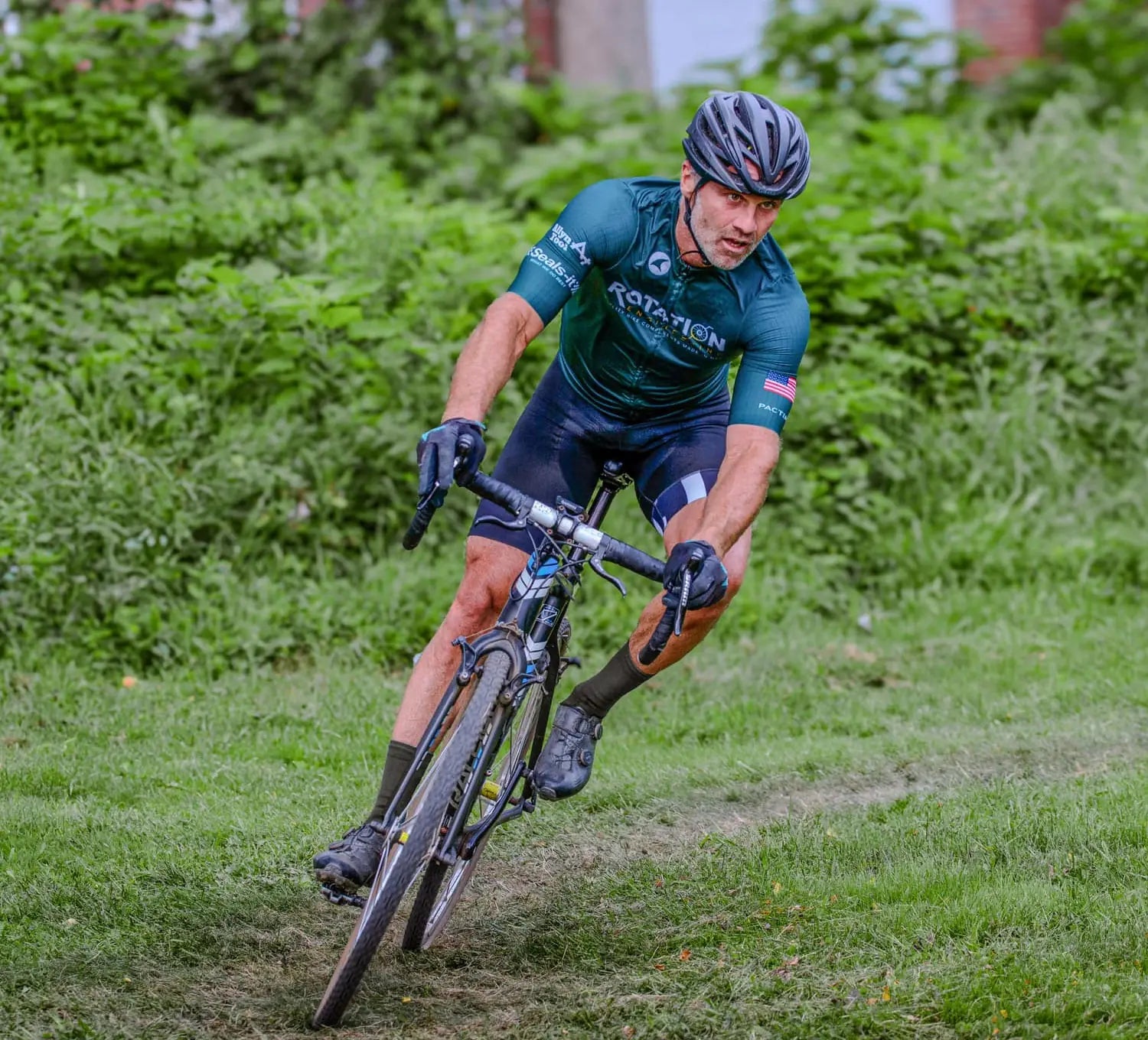 A cyclist in a green jersey and helmet leans into a curve on a grassy path, focused and determined. Lush green foliage surrounds the path, creating a natural setting.