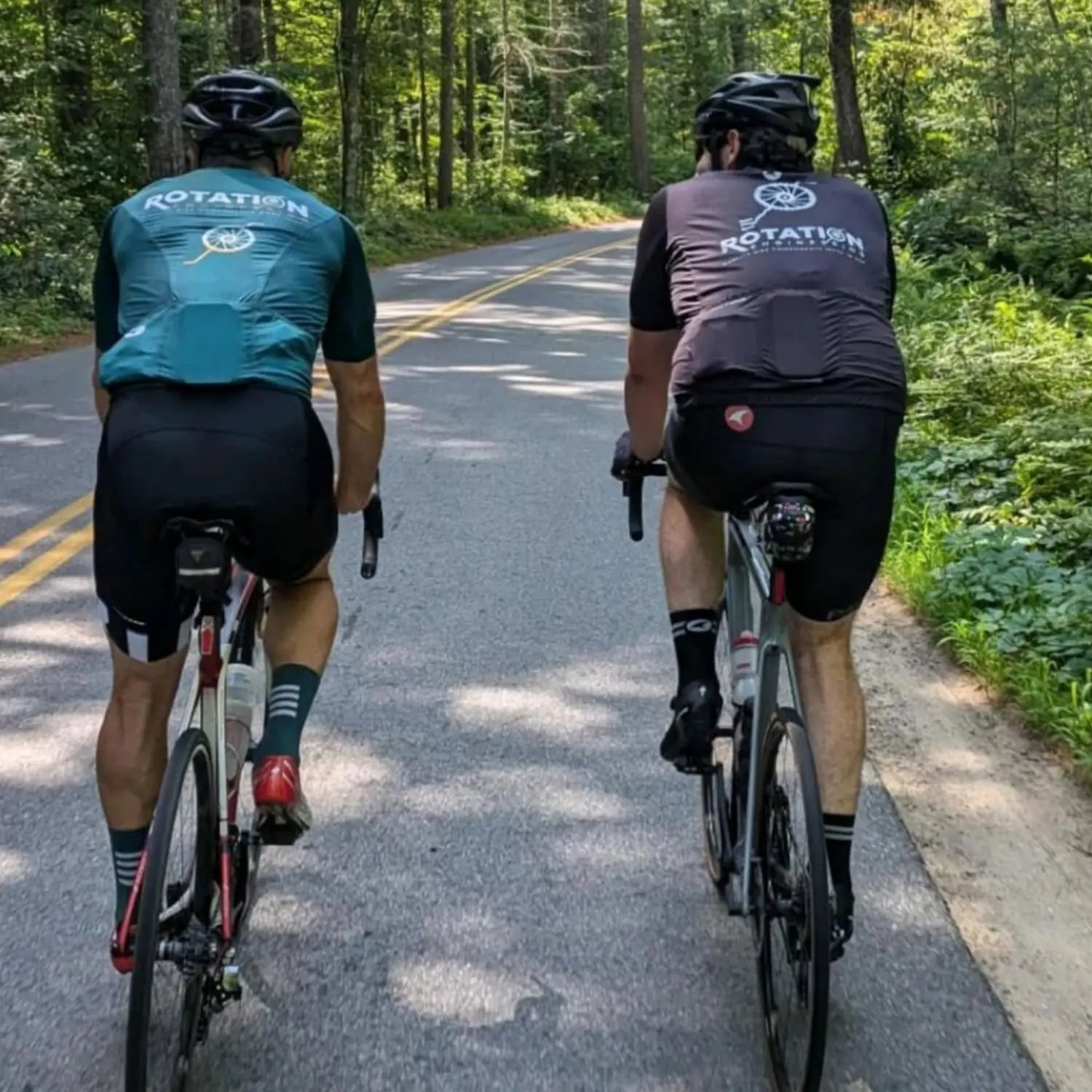Two cyclists wearing helmets and cycling gear ride on a forest road. Sunlight filters through trees, casting shadows. The mood is tranquil and focused.