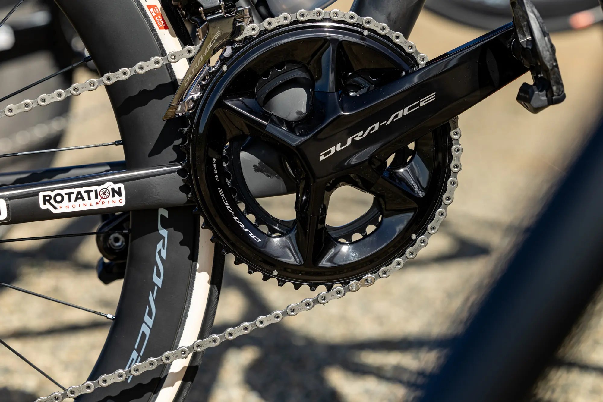 Close-up of a bicycle's black Dura-Ace crankset and chain, with the branding visible. The bike has sleek, aerodynamic features, evoking speed.