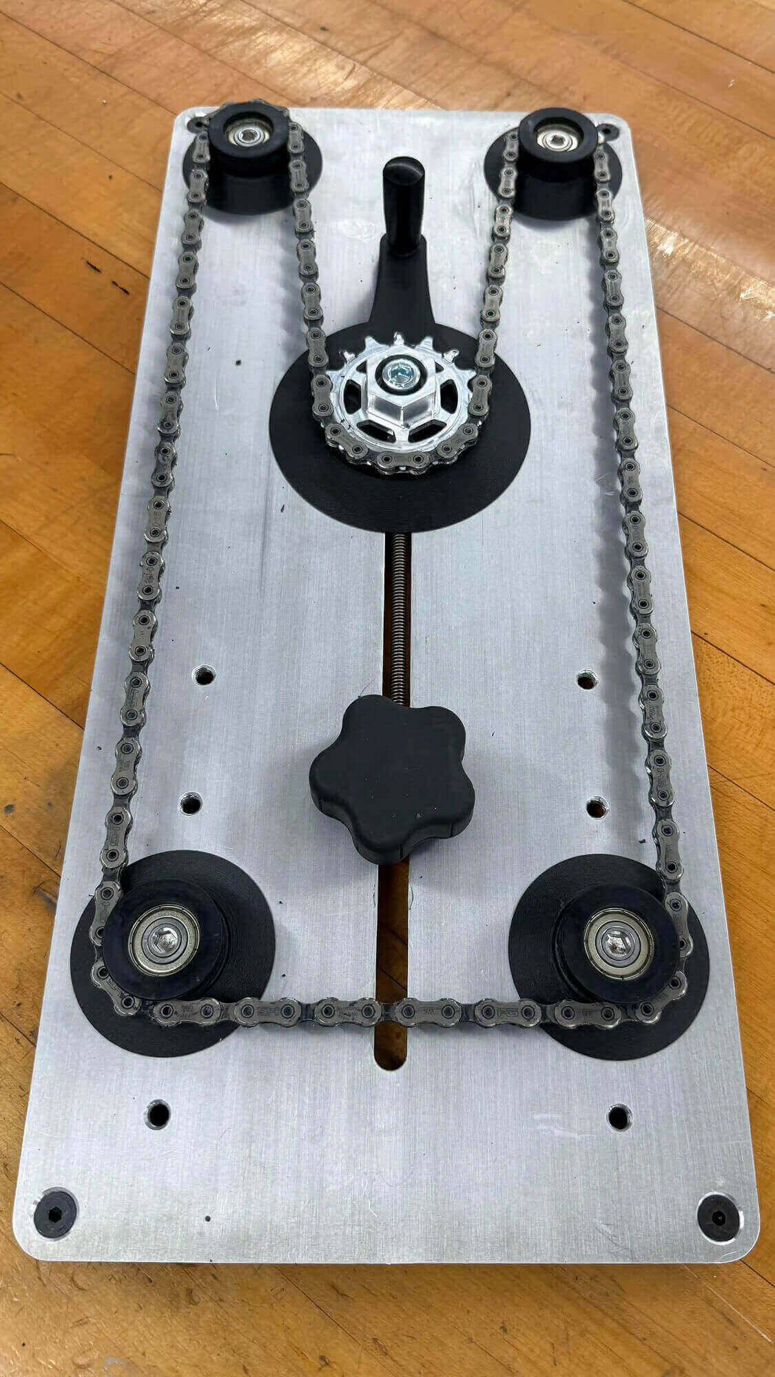 Close-up of a mechanical device on a wooden table, featuring a metal base, roller chain, gears, and a central knob, conveying an industrial feel.