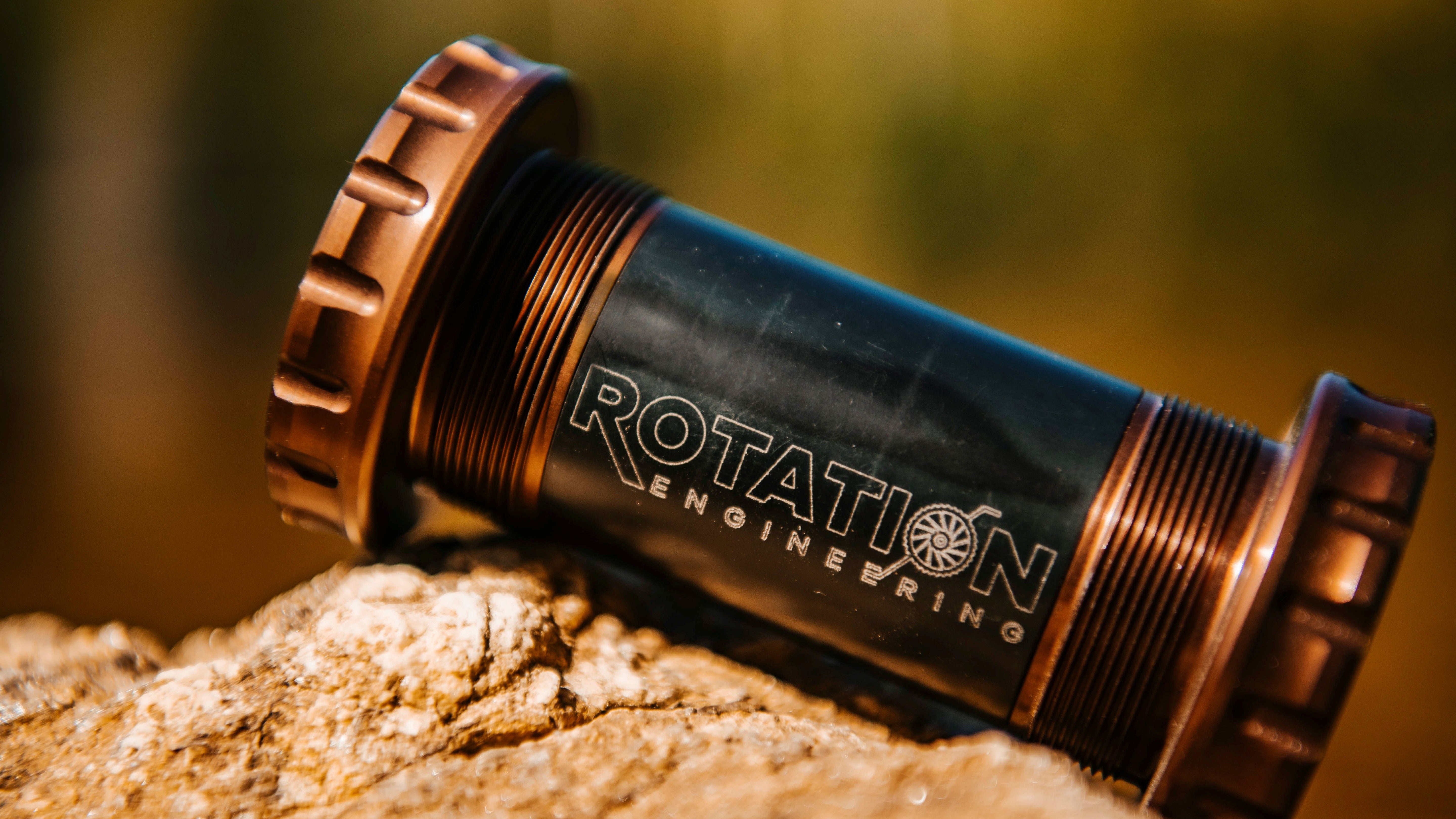 Close-up of a cylindrical bike part labeled "Rotation Engineering," resting on a rough rock surface. Warm, golden lighting enhances its metallic finish.