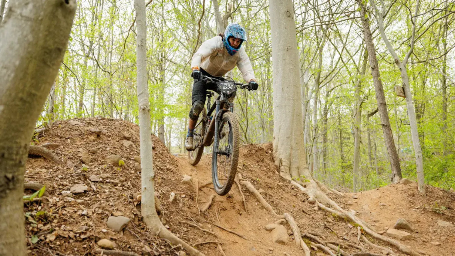 Mountain biker in a blue helmet navigates a dirt trail with exposed roots in a forest. The scene is energetic and adventurous, set in early spring.